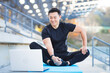 © Liubomir - handsome asian fitness coach holds workout classes online using video call webcam and laptop. sitting outside in a modern stadium. Pilates training or stretching. Physical exercises. Records Lessons
