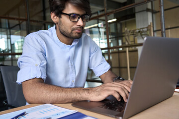 Wall Mural - Young smart Indian business man employee wearing glasses using computer working online in office. Ethnic guy entrepreneur or manager typing on laptop analyzing digital data technology at workplace.