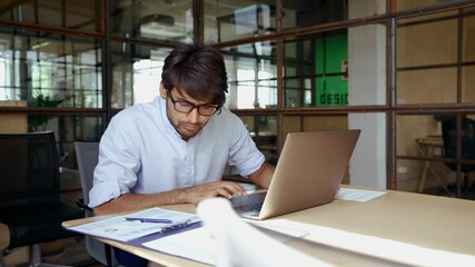 Wall Mural - Young smart Indian business man employee wearing glasses using computer working online in office. Ethnic guy entrepreneur or manager typing on laptop analyzing digital data technology at workplace.