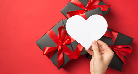 Wall Mural - First person top view photo of hand holding white paper heart over stack of gift boxes on isolated red background with blank space