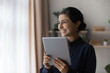 © fizkes - Dreamy smiling young Indian woman looking in distance, holding digital tablet in hands, feeling motivated getting message with pleasant news, enjoying online games or funny photo video content.