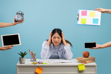 Deadline concept. Irritated asian lady stressed by a lot of work, sitting at the desk over blue studio background