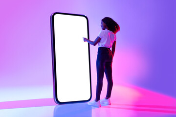 Wall Mural - Young black woman touching screen of giant smartphone in neon light, mockup for new mobile app or website