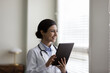 Smiling dreamy young Indian woman doctor physician in white uniform with stethoscope holding tablet, looking in distance, planning, visualizing, happy therapist practitioner consulting patient online