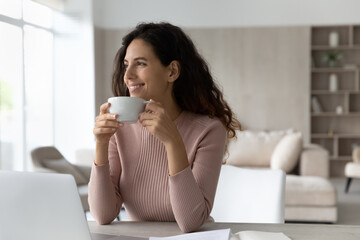 Smiling dreamy businesswoman drinking tea or coffee at workplace, happy confident woman looking to aside, holding white cup of hot beverage, enjoying break, visualizing new opportunities, future