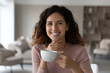 © fizkes - Head shot portrait smiling beautiful hispanic woman holding white cup of tea or coffee, leisure time with hot beverage at home, happy young female looking at camera, enjoying morning, breakfast