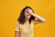 Portrait of woman with curly dark hair forgetting something, slapping forehead with palm and closing eyes isolated over yellow background