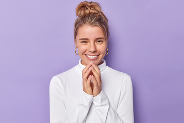 Wall Mural - Horizontal shot of pretty cheerful young European woman smiles gently steepls fingers and has intention to do something wears casual white turtleneck isolated over purple background schemes.
