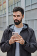 Photo of thoughtful handsome man dressed in black anorak uses mobile phone for chatting online downloads application for sport traning poses outdoors against blurred background. Modern lifestyle