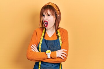 Wall Mural - Redhead young woman dressmaker designer wearing atelier apron angry and mad screaming frustrated and furious, shouting with anger. rage and aggressive concept.