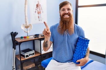 Sticker - Redhead man with long beard holding foam roller at medical clinic smiling happy pointing with hand and finger to the side