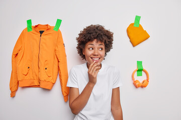 Wall Mural - Glad curly haired young woman smiles happily notices something pleasant away dressed in casual t shirt poses against white background with orange jacket hat and headphones plastered to wall.