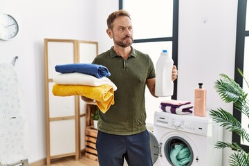 Wall Mural - Middle age man holding clean laundry and detergent bottle smiling looking to the side and staring away thinking.