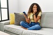 © Krakenimages.com - Beautiful hispanic woman sitting on the sofa at home using smartphone looking stressed and nervous with hands on mouth biting nails. anxiety problem.