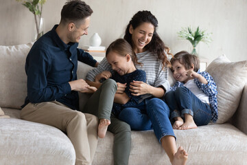 Laughing 30s couple tickling little preschool kids seated on sofa in living room. Hispanic parents enjoy playtime with adorable son and cute daughter. Games of happy family at home, love, fun concept