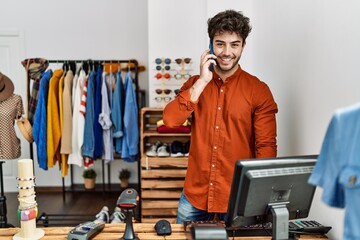 Wall Mural - Young hispanic shopkeeper man smiling happy talking on the smartphone working at clothing store.