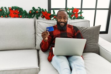 Sticker - Young african american man using laptop and credit card sitting by christmas decor at home