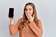 Young blonde woman holding smartphone showing screen covering mouth with hand, shocked and afraid for mistake. surprised expression