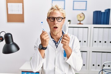 Poster - Young blond man working at dentist clinic holding electric toothbrush and teethbrush in shock face, looking skeptical and sarcastic, surprised with open mouth