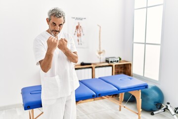 Wall Mural - Middle age hispanic therapist man working at pain recovery clinic ready to fight with fist defense gesture, angry and upset face, afraid of problem