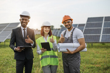 Wall Mural - Caucasian woman and african man standing with indian engineer among solar panels and looking at camera. People holding digital tablet, clipboard and drawings.