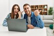 © Krakenimages.com - Young caucasian couple working using computer laptop at home pointing thumb up to the side smiling happy with open mouth
