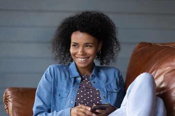 Wall Mural - Dreamy smiling attractive young African American woman holding cellphone in hands, thinking message or email with pleasant news, chatting communicating in social network resting on cozy sofa at home.