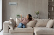 © fizkes - Smiling woman using air conditioner remote controller, relaxing on comfortable couch in living room alone, positive beautiful young female switching temperature on climate control system at home