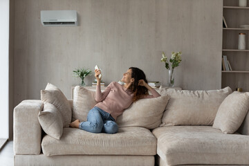 Smiling woman using air conditioner remote controller, relaxing on comfortable couch in living room alone, positive beautiful young female switching temperature on climate control system at home