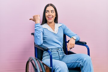 Poster - Beautiful woman with blue eyes sitting on wheelchair smiling with happy face looking and pointing to the side with thumb up.