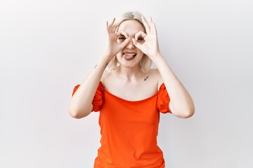 Poster - Young caucasian woman standing over isolated background doing ok gesture like binoculars sticking tongue out, eyes looking through fingers. crazy expression.