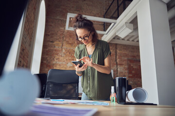 Joyful creative woman, interior designer or architect wearing glasses using smartphone and smiling, working on new project, standing in her office