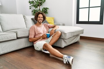 Sticker - Young hispanic man drinking coffee sitting on the floor at home.