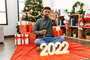 Poster - Young hispanic man with beard sitting by christmas tree celebrating 2022 new year with open hand doing stop sign with serious and confident expression, defense gesture