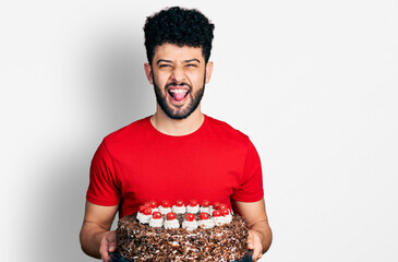 Sticker - Young arab man with beard celebrating birthday holding big chocolate cake sticking tongue out happy with funny expression.