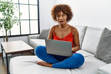 Sticker - Young african american woman sitting on the sofa at home using laptop afraid and shocked with surprise expression, fear and excited face.