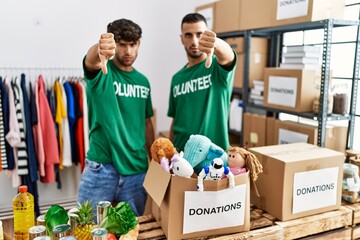 Poster - Young gay couple wearing volunteer t shirt at donations stand looking unhappy and angry showing rejection and negative with thumbs down gesture. bad expression.