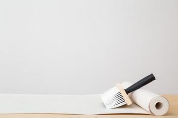 Brush and roll of wallpaper on wooden table at light gray wall background. Closeup. Repair work of home. Front view. Empty place for text.