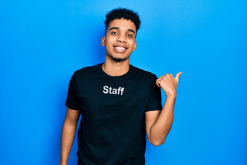 Young african american man wearing staff t shirt pointing to the back behind with hand and thumbs up, smiling confident