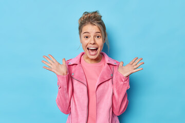 Canvas Print - Horizontal shot of amazed young European woman raises palms and exclaims loudly reacts on awesome present or surprise wears pink jacket isolated over blue studio wall. People and reactions concept