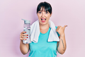 Poster - Young hispanic woman wearing sportswear holding water bottle pointing thumb up to the side smiling happy with open mouth