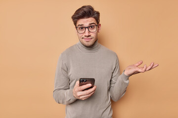 Confused questioned young man shrugs shoulders wears optical glasses and turtleneck holds mobile phone reads something strange browses newsfeed via social networks puzzled about online content