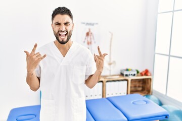 Sticker - Young handsome man with beard working at pain recovery clinic shouting with crazy expression doing rock symbol with hands up. music star. heavy concept.