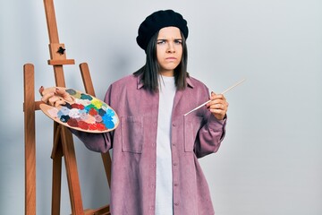 Canvas Print - Young brunette woman standing by empty easel stand holding palette skeptic and nervous, frowning upset because of problem. negative person.