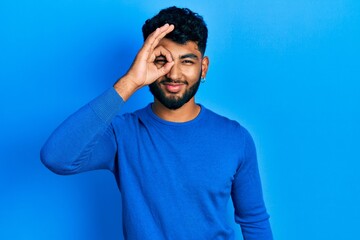 Poster - Arab man with beard wearing casual blue sweater doing ok gesture with hand smiling, eye looking through fingers with happy face.