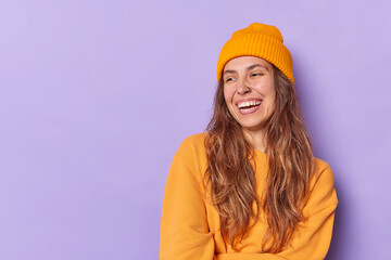 Wall Mural - Studio shot of happy teenage girl with toothy smile laughs joyfully has piercing in nose wears orange hat and sweatshirt isolated over purple background blank space for your promotional content