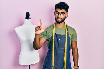 Wall Mural - Arab man with beard dressmaker designer wearing atelier apron smiling with an idea or question pointing finger up with happy face, number one