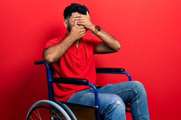 Wall Mural - Arab man with beard sitting on wheelchair covering eyes and mouth with hands, surprised and shocked. hiding emotion