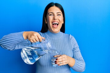 Poster - Beautiful brunette woman pouring water in glass smiling and laughing hard out loud because funny crazy joke.