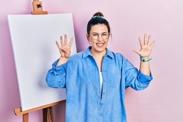 Poster - Young hispanic woman standing by painter easel stand showing and pointing up with fingers number nine while smiling confident and happy.